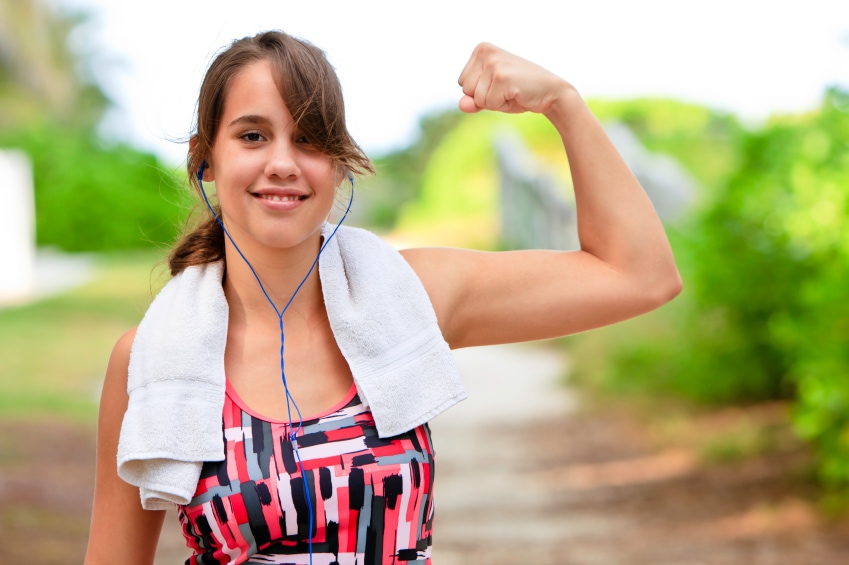 Smiling teenage female wearing sports clothing, towel around neck, and earphones flexing bicep