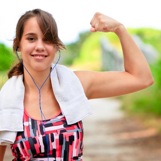 Smiling teenage female wearing sports clothing, towel around neck, and earphones flexing bicep